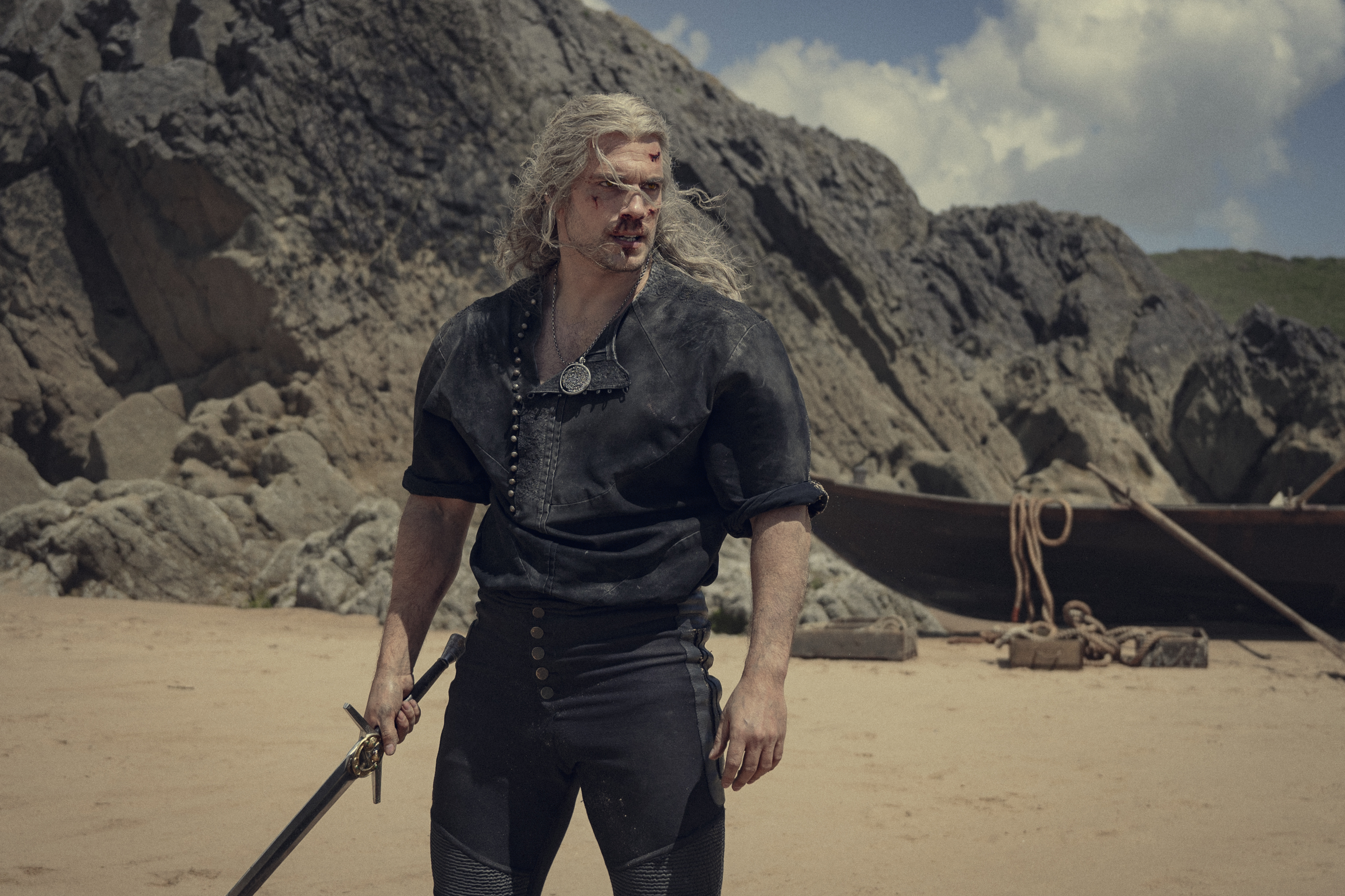 Geralt stands on a beach with a bloody nose and his sword held at his side in The Witcher season 3