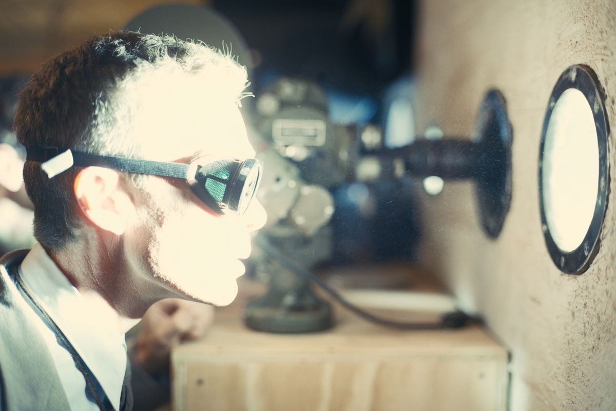 As J. Robert Oppenheimer in the film Oppenheimer, Cillian Murphy wears goggles and stares through a porthole at an explosion casting an intensely bright light on his face