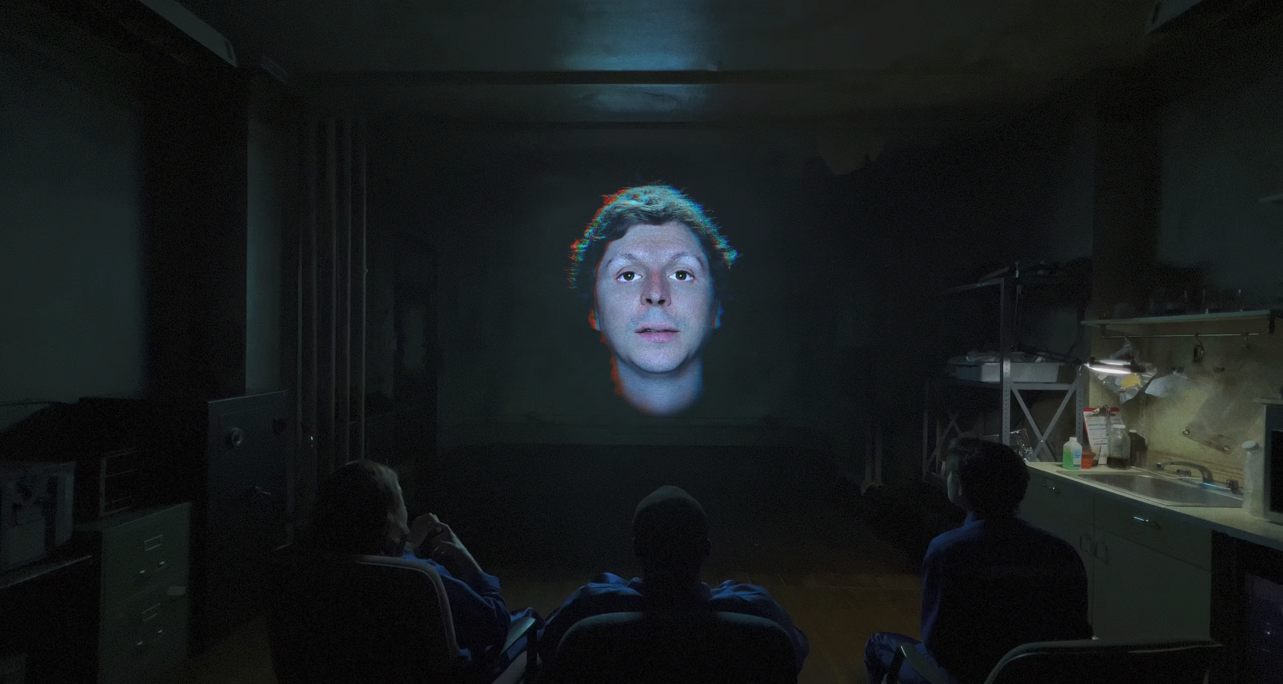 A disembodied Michael Cera head is projected on the wall in front of three people in a shabby living room in Command Z. 